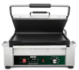 A stainless steel Waring Panini Grill with a partially open lid, square cast iron plates, and a control panel featuring a digital timer display, temperature knob, ready light, and power switch. The label reads Waring Tostato Supremo.