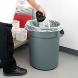 A person in black shoes and dark pants throws a small black trash bag into a heavy duty, 20 gallon Trash Can lined with a clear plastic bag. The can sits by a metal table with white containers and red cutting boards in a clean, indoor setting.