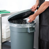 A person in a black apron lifts the Winco PTCL-20G lid of a large gray trash can lined with a clear plastic bag, about to throw away a crumpled item. Nearby, a metal cart with white containers and a green cutting board sits in the clean indoor space.