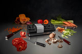A kitchen scene centers on an electric knife sharpener, surrounded by sliced and whole tomatoes, peppers, carrots, mushrooms, celery, and squash. Three knives rest on a reflective countertop, with the sharpener’s grinding wheels subtly in view.