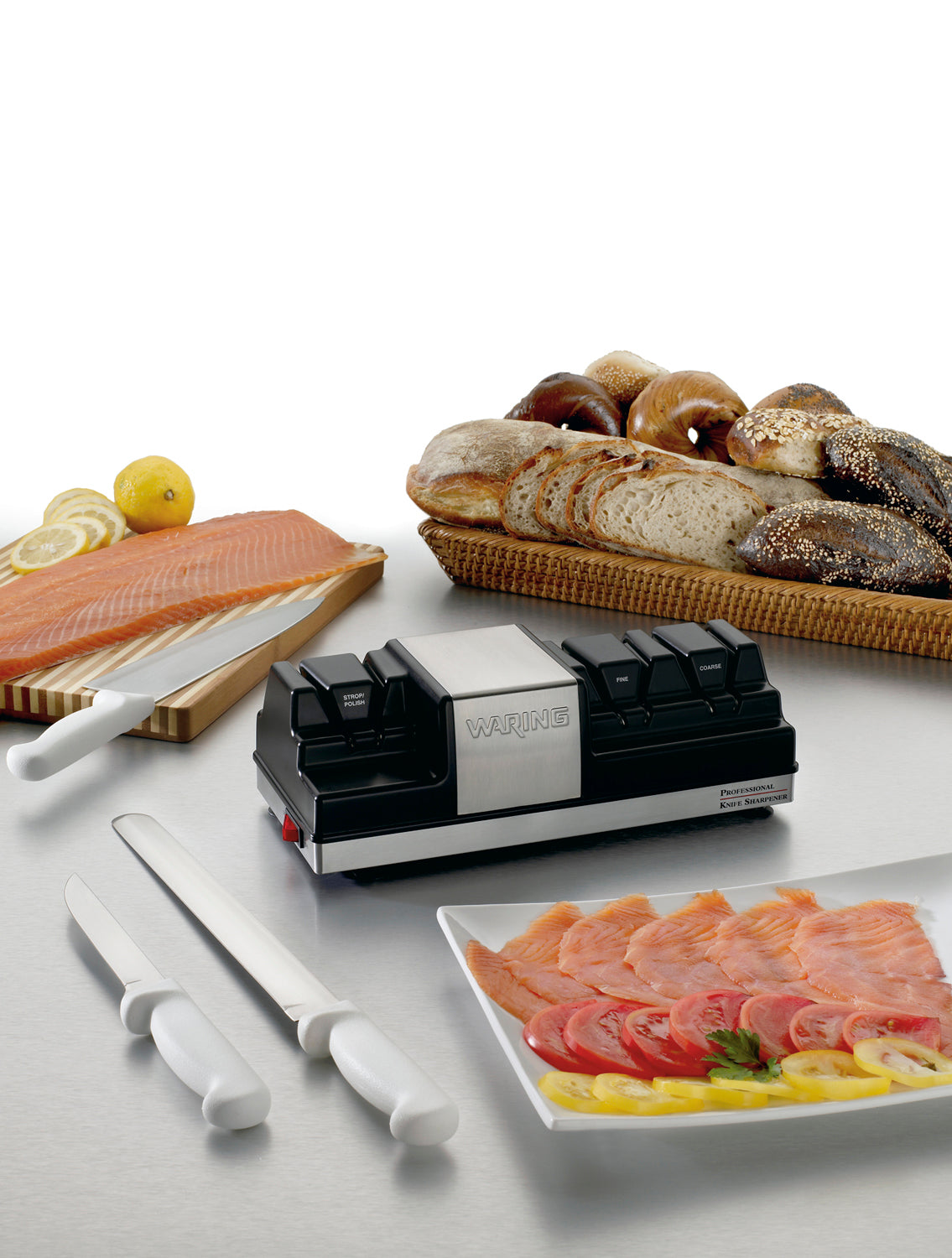 An electric Knife Sharpener sits on a kitchen counter. Nearby are knives, a cutting board with smoked salmon and lemon, a platter of sliced salmon with garnish, and a basket of assorted bagels. The background is plain white.