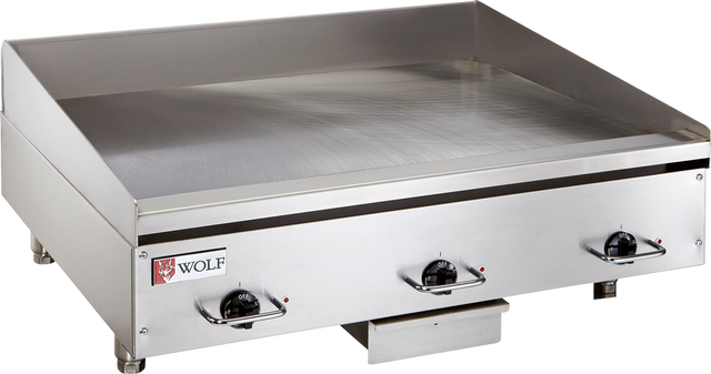 A commercial electric griddle featuring three black control knobs, raised side and back splashes, and a small Wolf brand logo on the front left corner. This stainless steel griddle offers a flat cooking surface with a grease tray beneath the front edge.