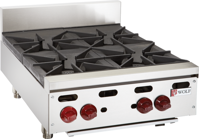 A stainless steel commercial hotplate with four black cast iron burners, red control knobs, and metal legs. The Wolf logo is visible on the front panel. This professional kitchen appliance features a raised backguard and a modern, industrial design.