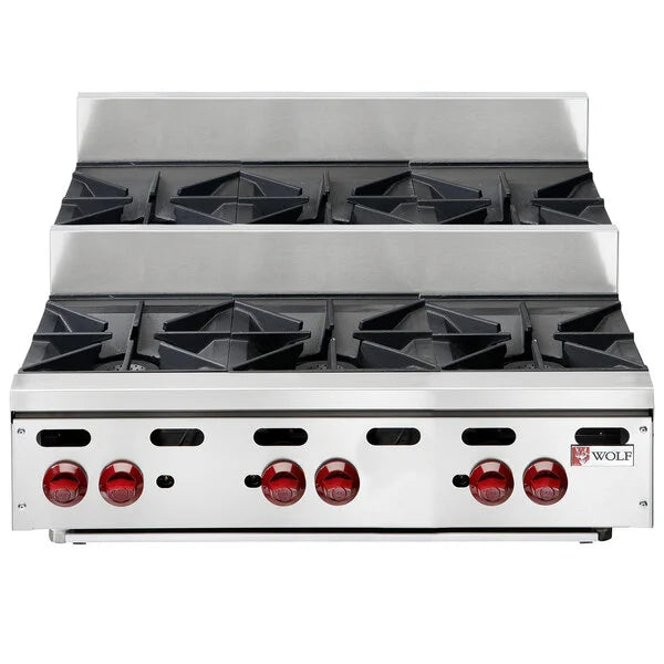 A stainless steel Wolf Achiever Hotplate stovetop with six black cast iron burners and six red control knobs. This 36-inch hotplate features a raised back panel and a sleek, professional design found in commercial kitchens.