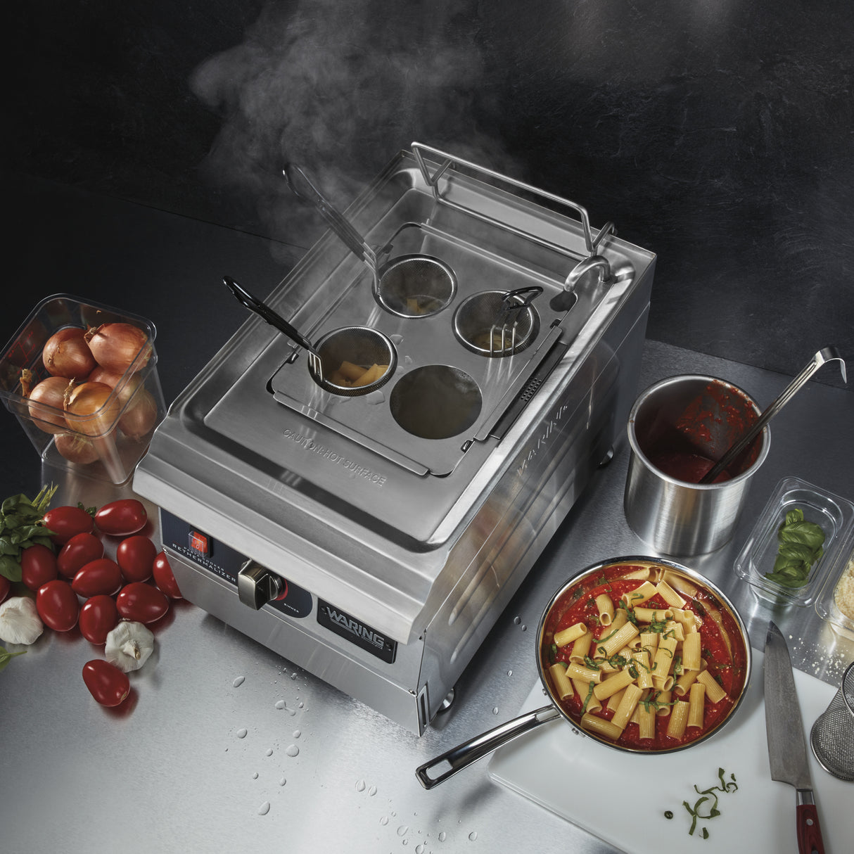 A Waring WPC100K electric pasta cooker with four baskets steams on a stainless steel counter, surrounded by rigatoni in tomato sauce, a pan of tomato sauce with basil, cherry tomatoes, eggs, grated cheese, and chopped green herbs.