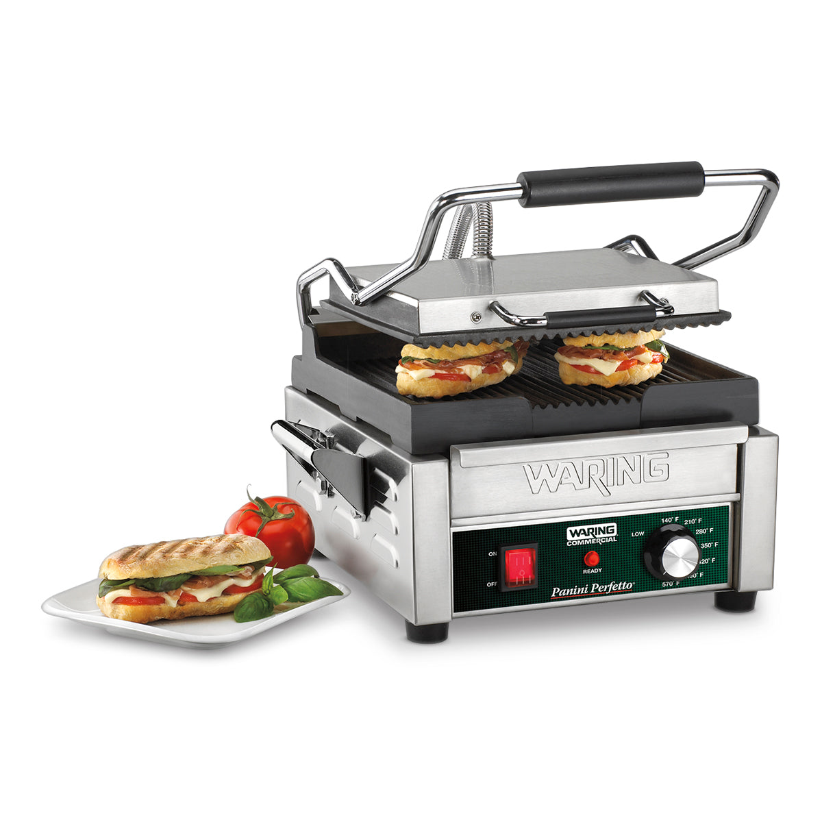 A compact panini grill with ribbed cast iron plates grills two sandwiches with melted cheese and tomato, while a finished grilled panini sits on a white plate. The machine features an adjustable thermostat and a control knob on the front.
