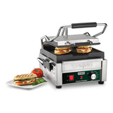A compact panini grill with ribbed cast iron plates grills two sandwiches with melted cheese and tomato, while a finished grilled panini sits on a white plate. The machine features an adjustable thermostat and a control knob on the front.