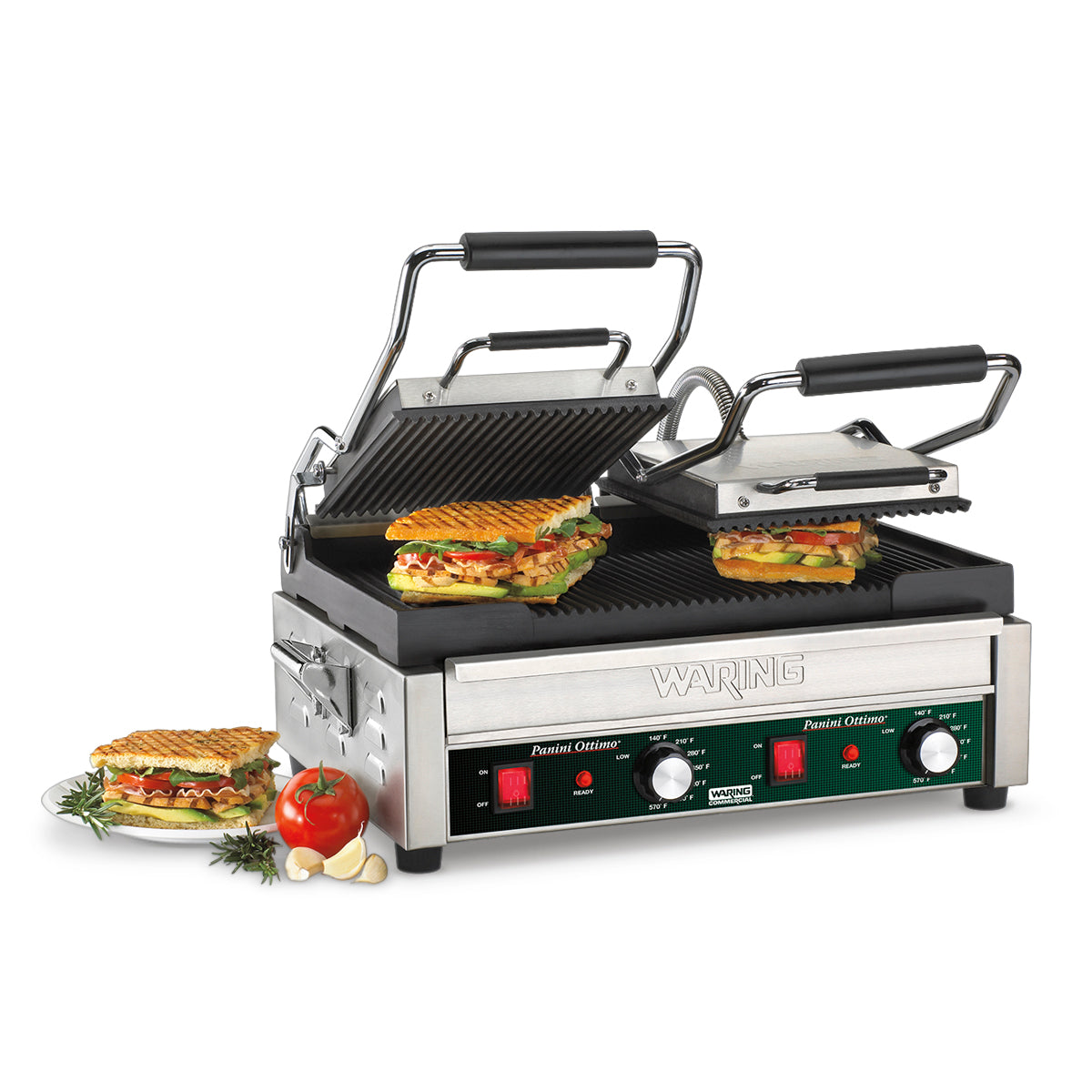 A stainless steel Waring electric panini grill with two ribbed grilling plates cooks two panini sandwiches. Finished paninis rest on a glass plate with tomato, garlic, and rosemary garnish. The grill features control dials and red/green lights.