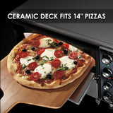 A freshly baked pizza with mozzarella, cherry tomatoes, olives, and basil is being placed into a black electric countertop pizza oven with a wooden peel. Text at the top reads, CERAMIC DECK FITS 14 PIZZAS. Oven controls are visible on the right.