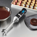 An immersion blender with a stainless steel shaft rests on a countertop beside chocolate sauce, colorful macarons, and a sea-salt-topped dessert, suggesting a professional or home kitchen setting.
