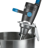 A close-up of a Big Stix immersion blender with a black and blue handle attached to the side of a large stainless steel mixing bowl using a sturdy bowl clamp. The blender features a secure mounting bracket and visible vent slots near the handle.