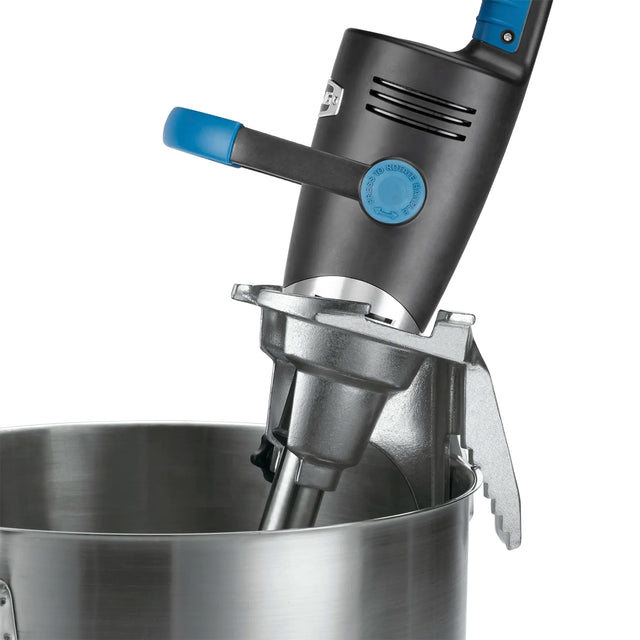 A close-up of a Big Stix immersion blender with a black and blue handle attached to the side of a large stainless steel mixing bowl using a sturdy bowl clamp. The blender features a secure mounting bracket and visible vent slots near the handle.