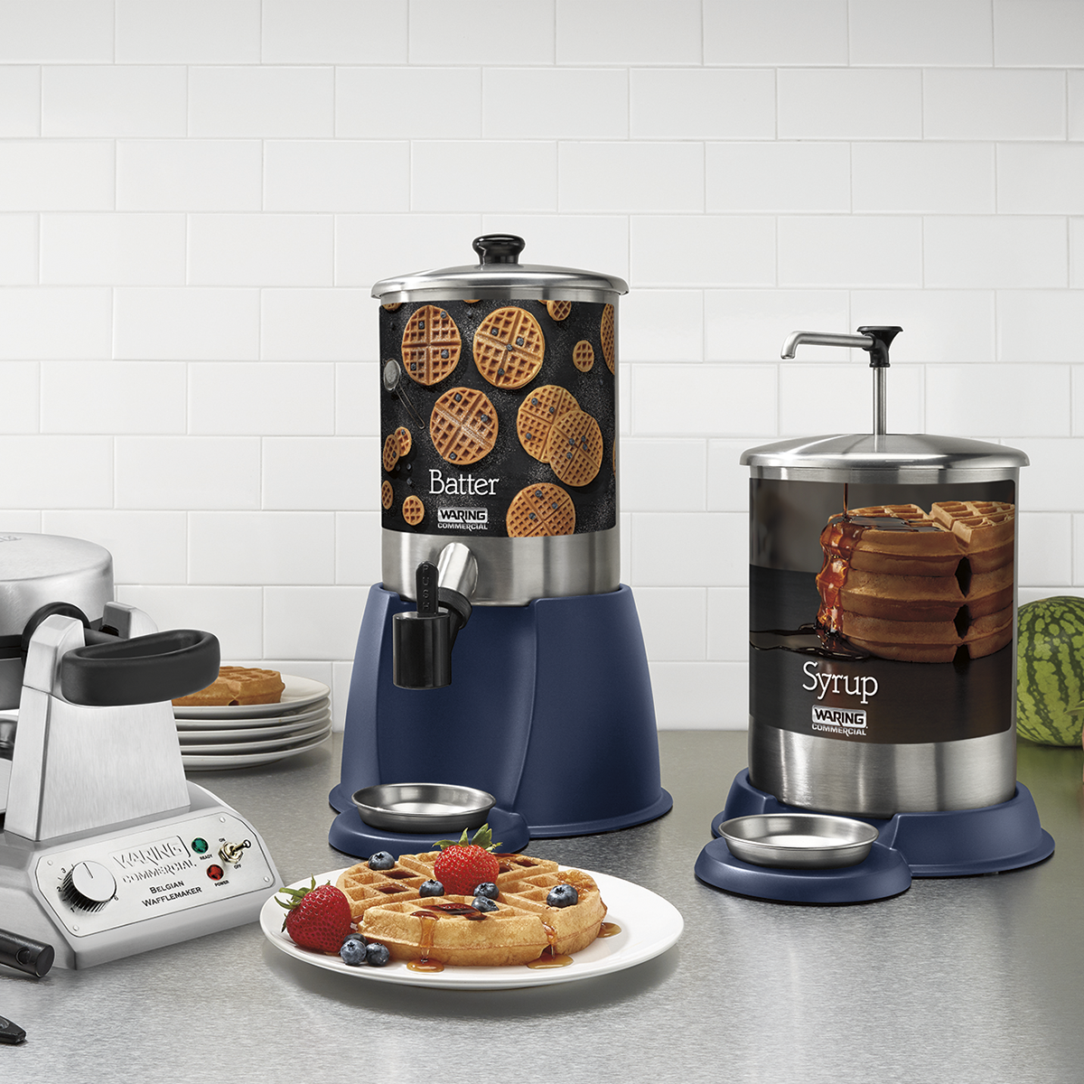 A kitchen counter displays a stainless steel 1-gallon syrup dispenser and a waffle batter dispenser, both with blue bases and waffle images. A waffle maker and a plate with waffles, strawberries, blueberries, and whipped cream are also shown.