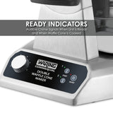 Close-up of a stainless steel Waring Commercial Double Waffle Cone Maker with non-stick plates. The display shows numbered lights, a dial, and start/stop buttons. Text above: Ready Indicators—Audible chime signals when unit is ready and cone is cooked.