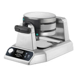 A stainless steel Waring commercial double Waffle Cone Maker with two round non-stick plates, a black handle, control buttons, and indicator lights on a digital front panel. The machine rests on a small metal tray to catch drips.
