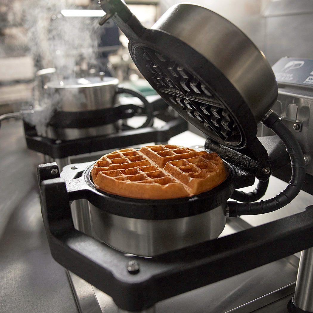 A golden brown waffle sits inside an open heavy-duty Belgian Waffle Maker with cast-iron plates on a stainless steel counter, steam rising from the hot plates. Another waffle maker is visible in the background, slightly out of focus.