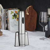 A rechargeable wine opener with a sleek stainless steel design rests on a marble countertop, accompanied by a glass of red wine, a bottle, cookbooks, and wooden cutting boards with rope handles leaning against the backsplash.